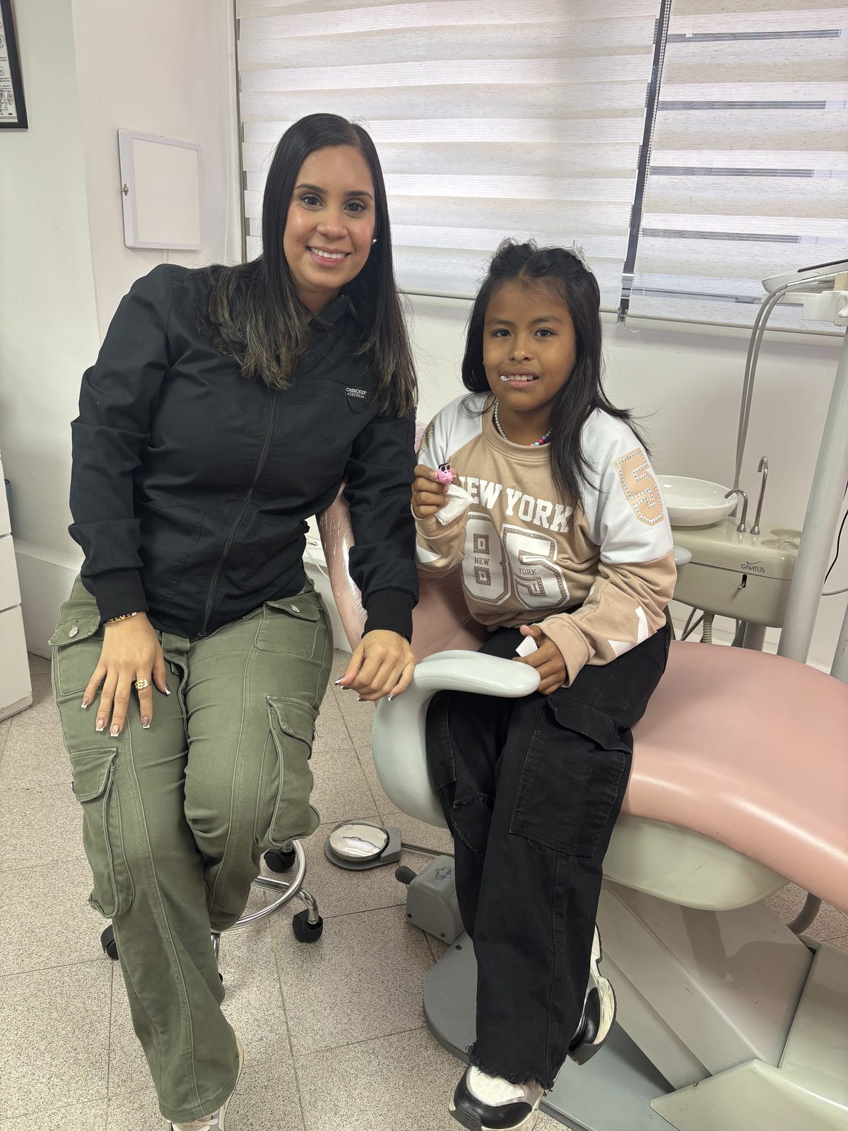 La Dra. Katherin Díaz sentada junto a una niña paciente en el sillón dental, ambas sonriendo